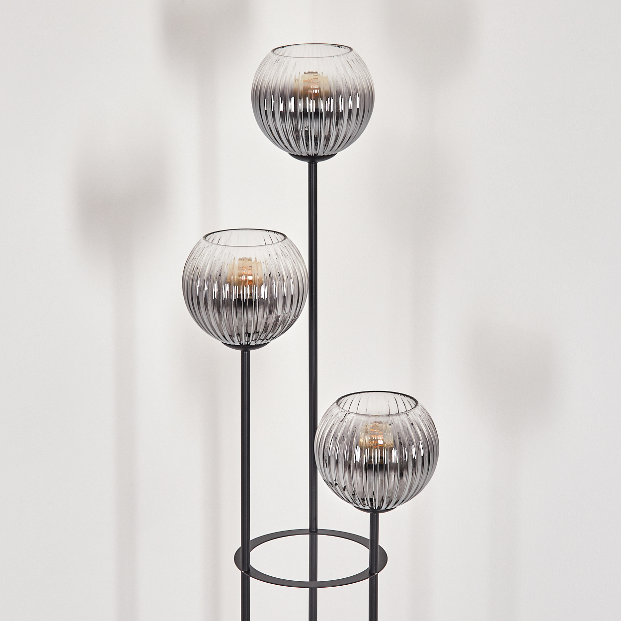 Eine moderne Stehlampe mit drei kugelförmigen, geriffelten Glasschirmen. Die Lampe hat ein schwarzes Metallgestell. Der Hintergrund ist eine weiße Wand.