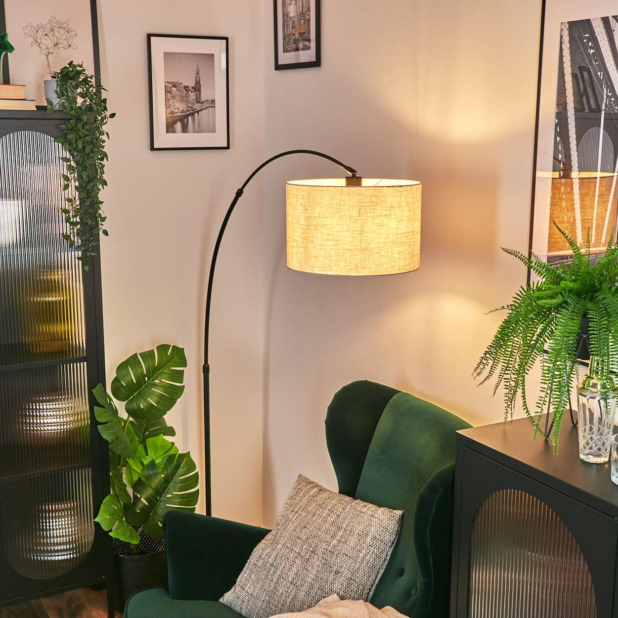 Ein Raum mit einem grünen Sessel, einer Stehlampe und einem Schrank. Es gibt Pflanzen, Bilder und ein Kissen. Die Lampe ist an.