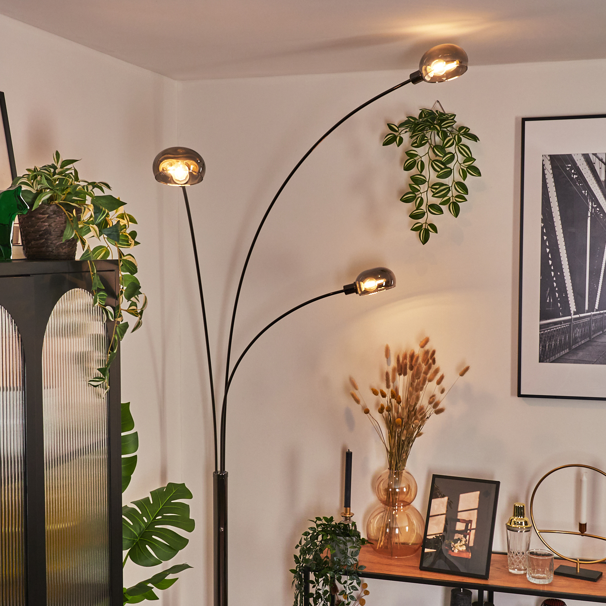 Ein modernes Wohnzimmer mit einer schwarzen Stehlampe, Pflanzen und einem gerahmten Bild. Die Lampe hat drei Lichter.