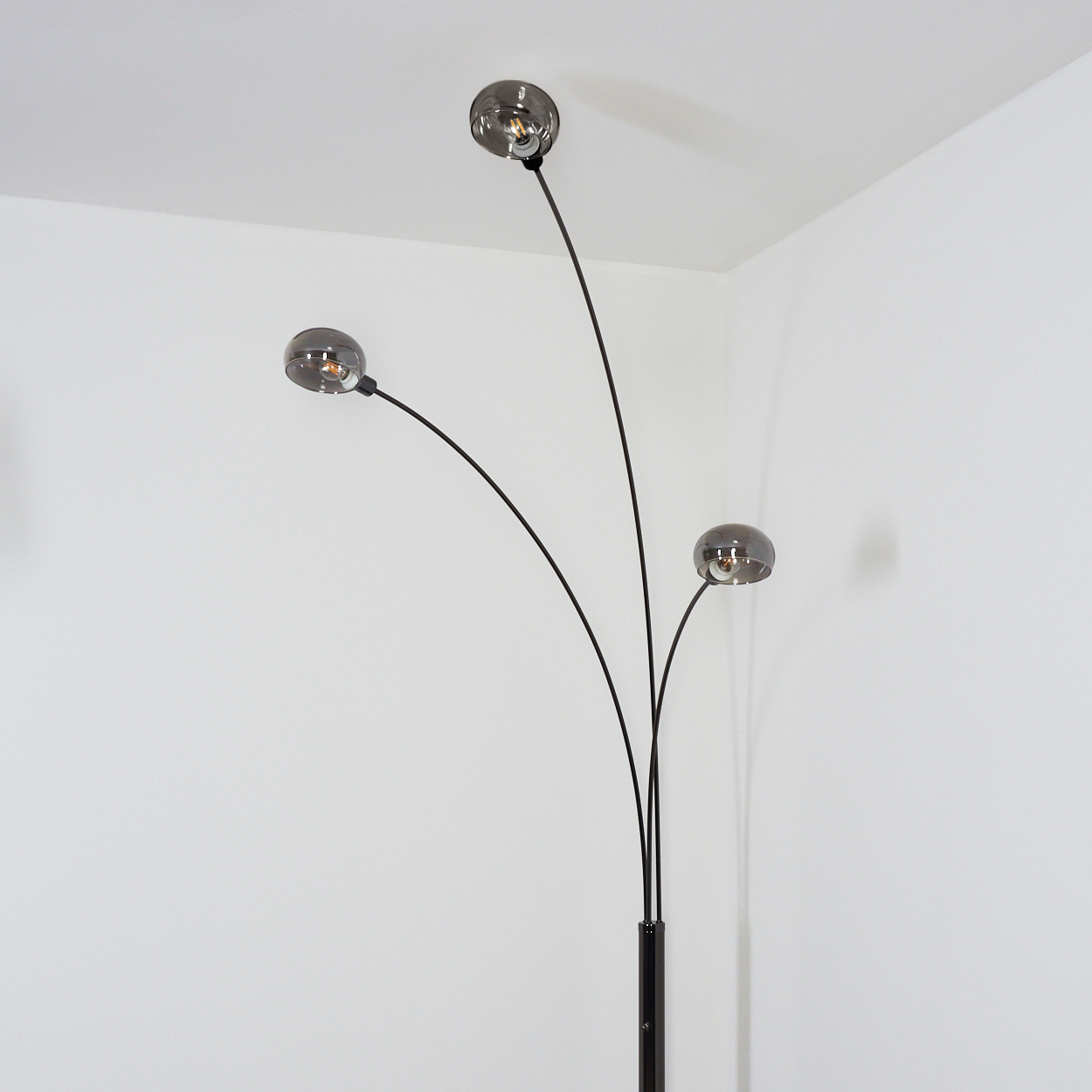 Eine moderne Stehlampe mit drei gebogenen Armen, die jeweils einen rauchigen Glaskugelschirm tragen. Die Lampe ist schwarz vor einer weißen Wand.