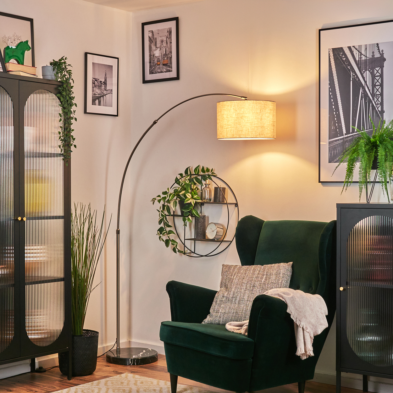 Ein Wohnzimmer mit grünem Sessel, Stehlampe und schwarzem Schrank. Bilder und Pflanzen schmücken die Wand.
