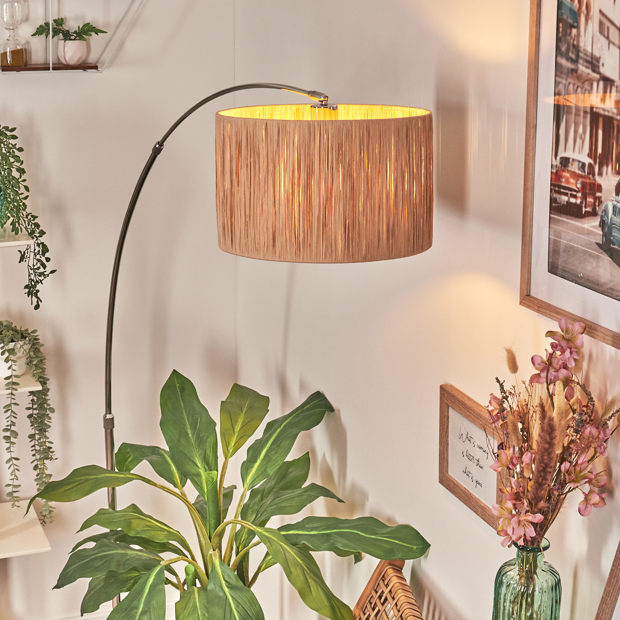 Eine Stehlampe mit beigem Schirm steht in einem Raum mit Pflanzen, Bildern und einem Korbstuhl. Die Lampe hat einen gebogenen Metallarm.