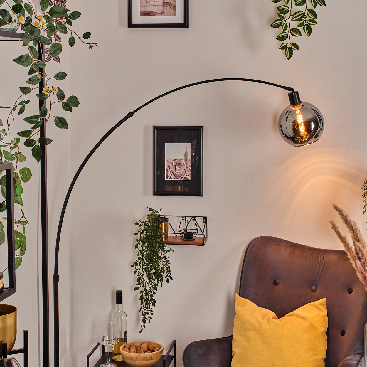 Ein modernes Wohnzimmer mit einer schwarzen Bogenlampe, einem grauen Sessel und einem gelben Kissen. Bilder und Pflanzen schmücken die Wand.
