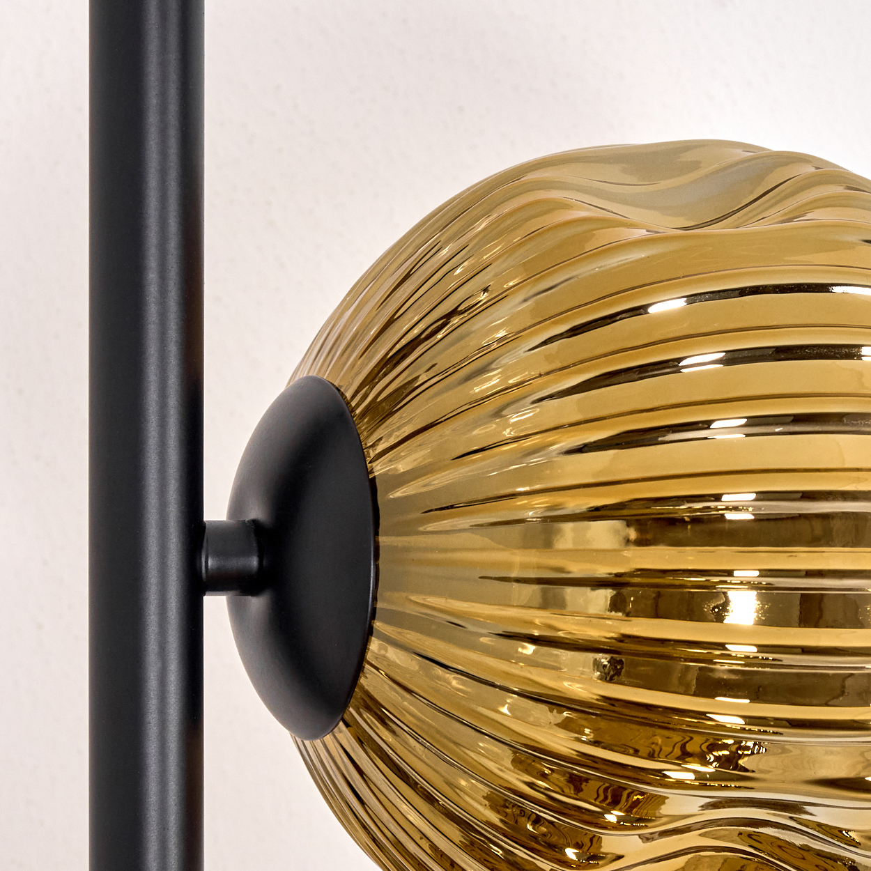 Nahaufnahme einer modernen Lampe. Sie hat einen schwarzen Metallrahmen und eine goldene, geriffelte Glaskugel. Der Hintergrund ist eine weiße Wand.
