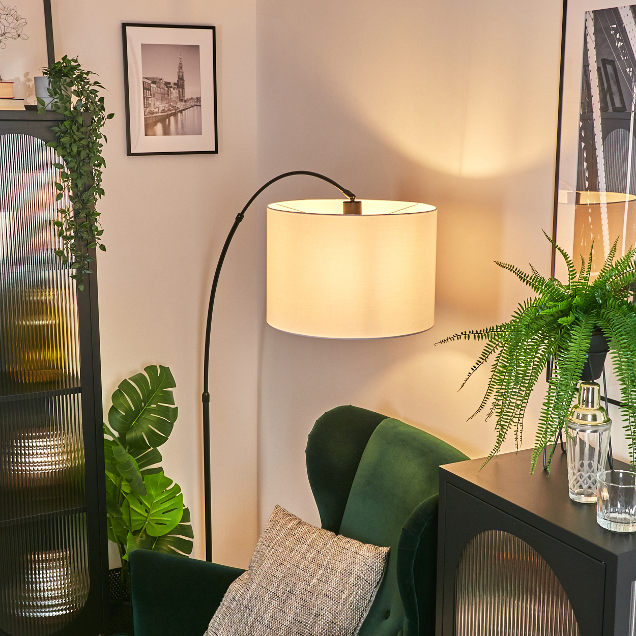 Ecke eines Wohnzimmers mit grünem Sessel, Lampe, schwarzem Schrank und gerahmten Bildern. Pflanzen verleihen der Szene eine natürliche Note.