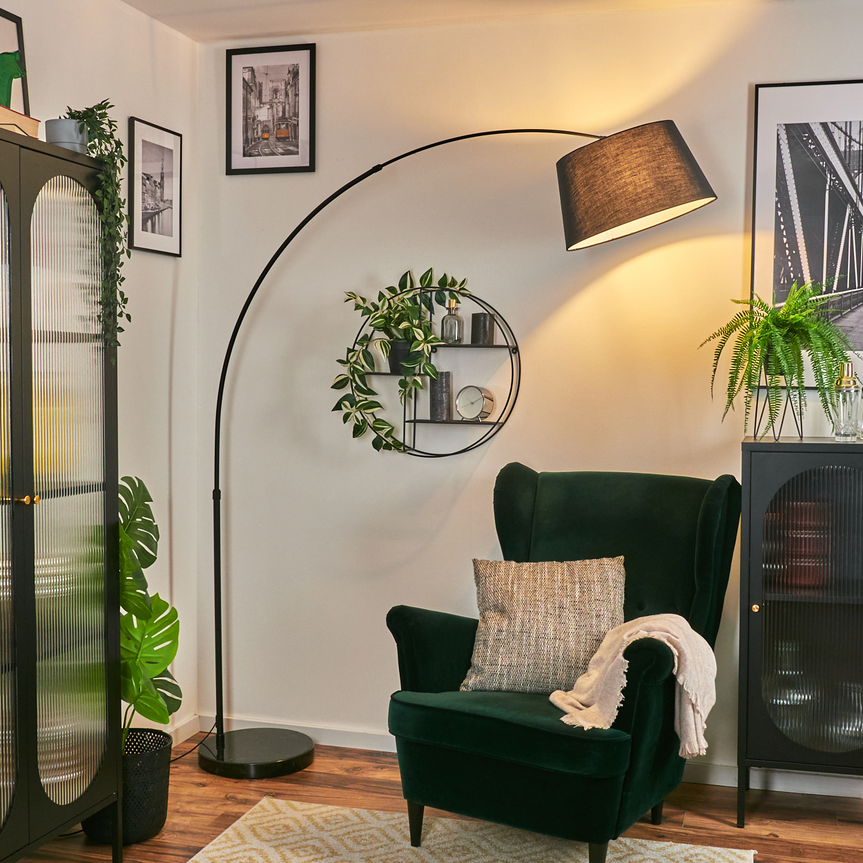Ein grüner Sessel mit Kissen und Decke steht unter einer schwarzen Bogenlampe. Es gibt zwei schwarze Schränke und gerahmte Bilder an der Wand.