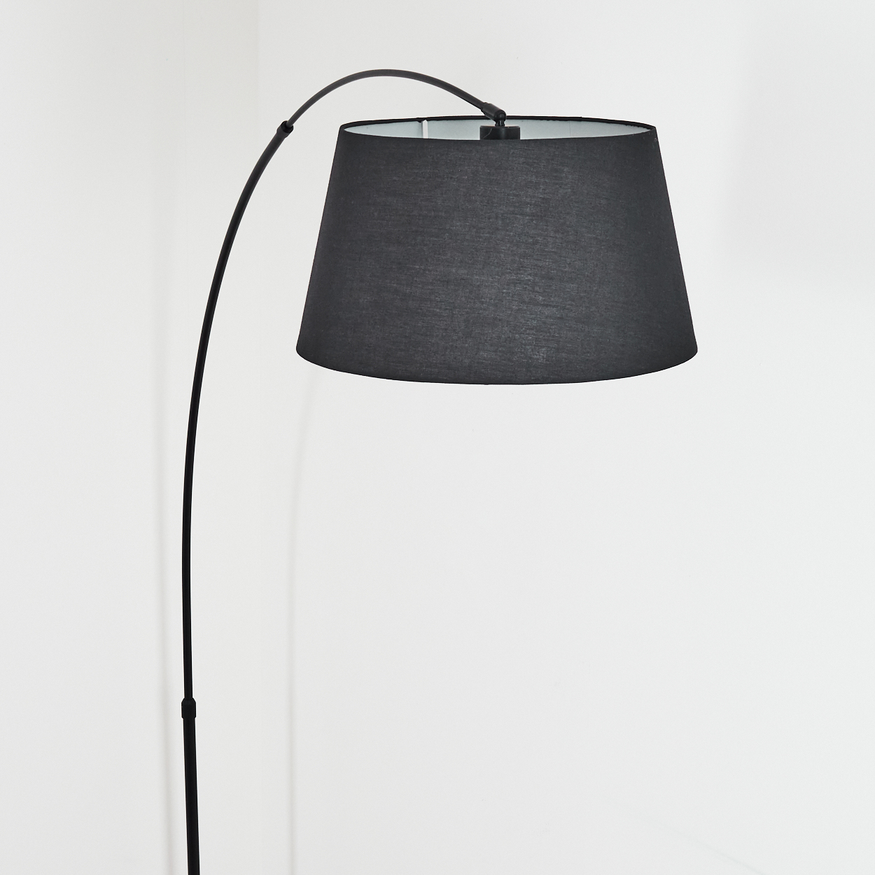 Eine schwarze Bogen-Stehlampe mit großem Schirm steht vor einer weißen Wand. Die Lampe ist aus.