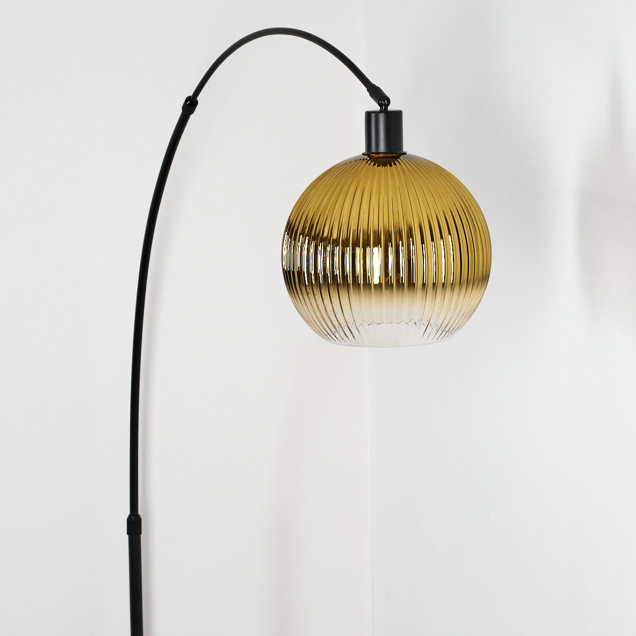 Eine moderne Lampe mit goldenem, geripptem Glasschirm hängt an einem schwarzen, gebogenen Arm. Der Hintergrund ist eine weiße Wand.