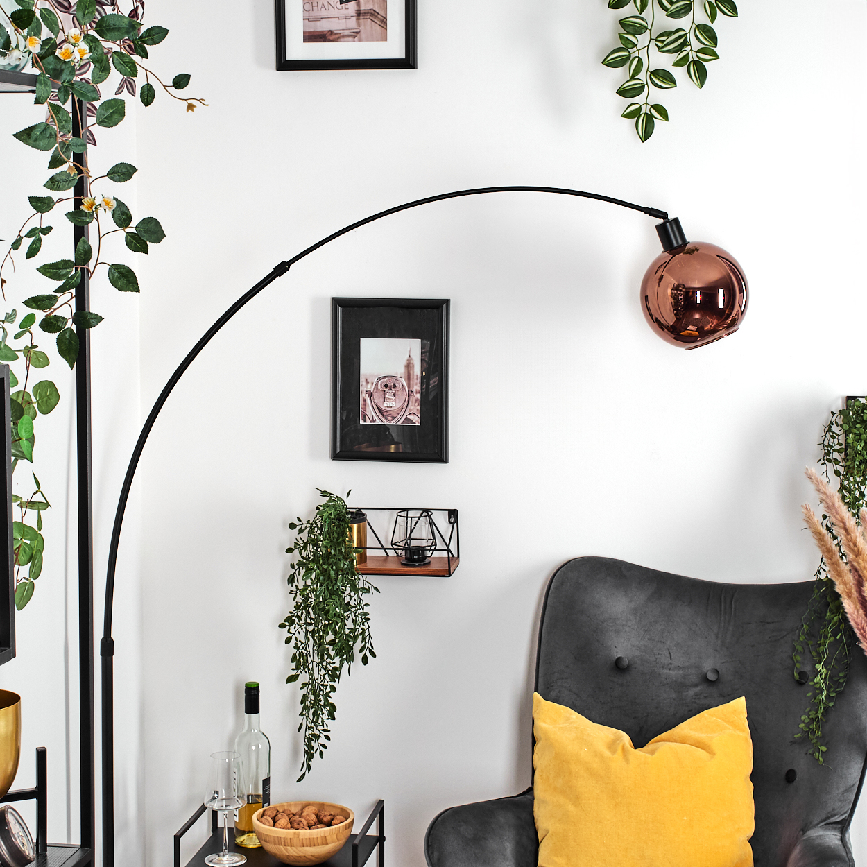 Ein Raum mit einer schwarzen Bogenlampe, Bildern, Hängepflanzen, einem grauen Sessel mit gelbem Kissen und einem Tisch.
