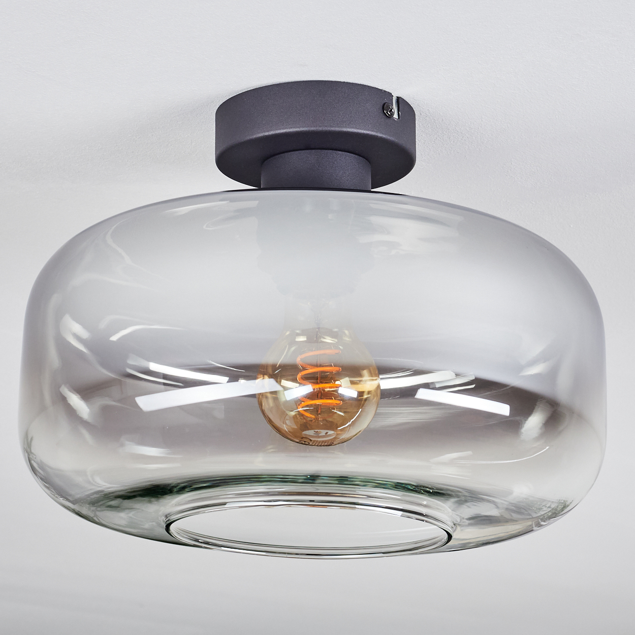 Eine moderne Deckenleuchte. Sie hat einen Rauchglasschirm mit einer Glühbirne. Die Basis ist dunkelgrau, an einer weißen Decke befestigt.