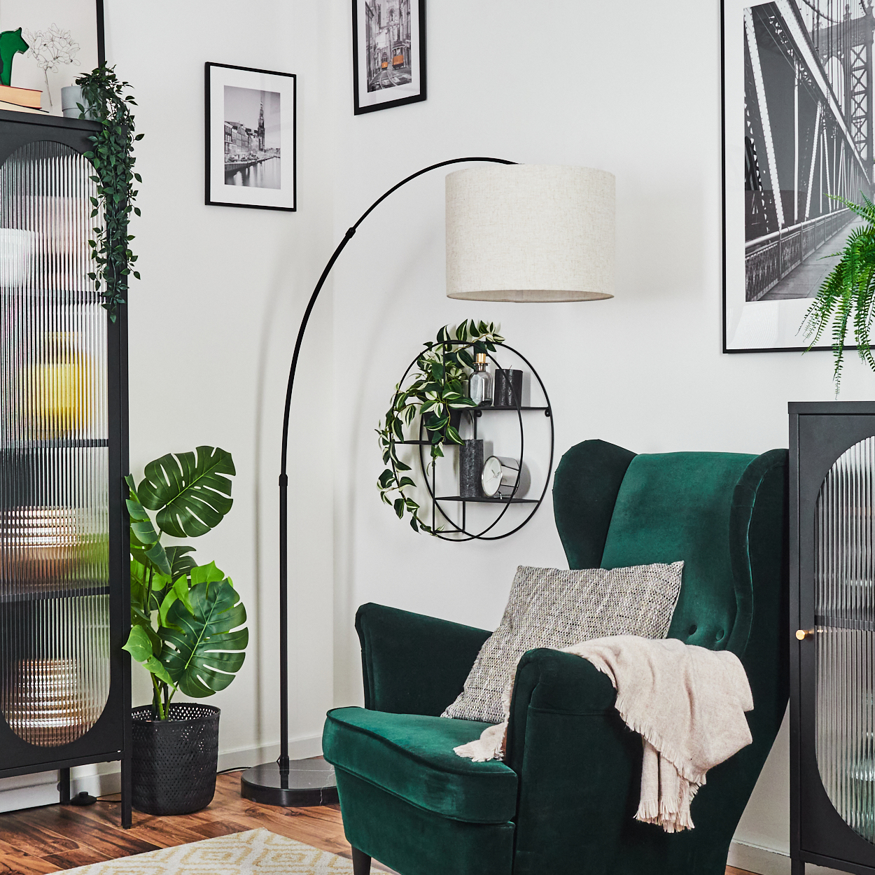 Ein grüner Sessel mit Decke und Kissen, eine schwarze Lampe, ein rundes Regal mit Pflanzen und gerahmte Bilder an einer weißen Wand.