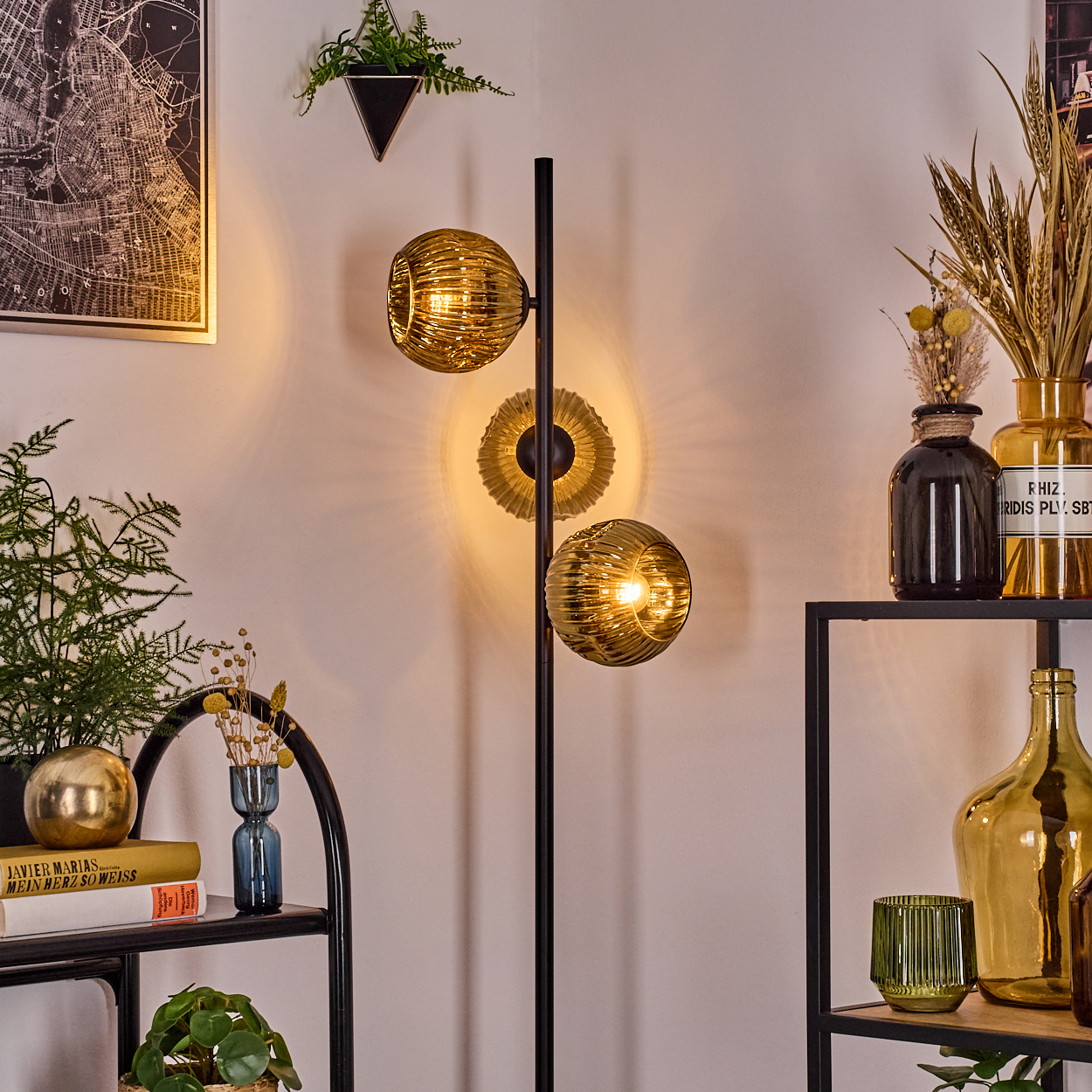 Ein Raum mit einer modernen Lampe, Regalen mit Vasen, Pflanzen und Büchern. Die Lampe hat drei bernsteinfarbene Glaskugeln.