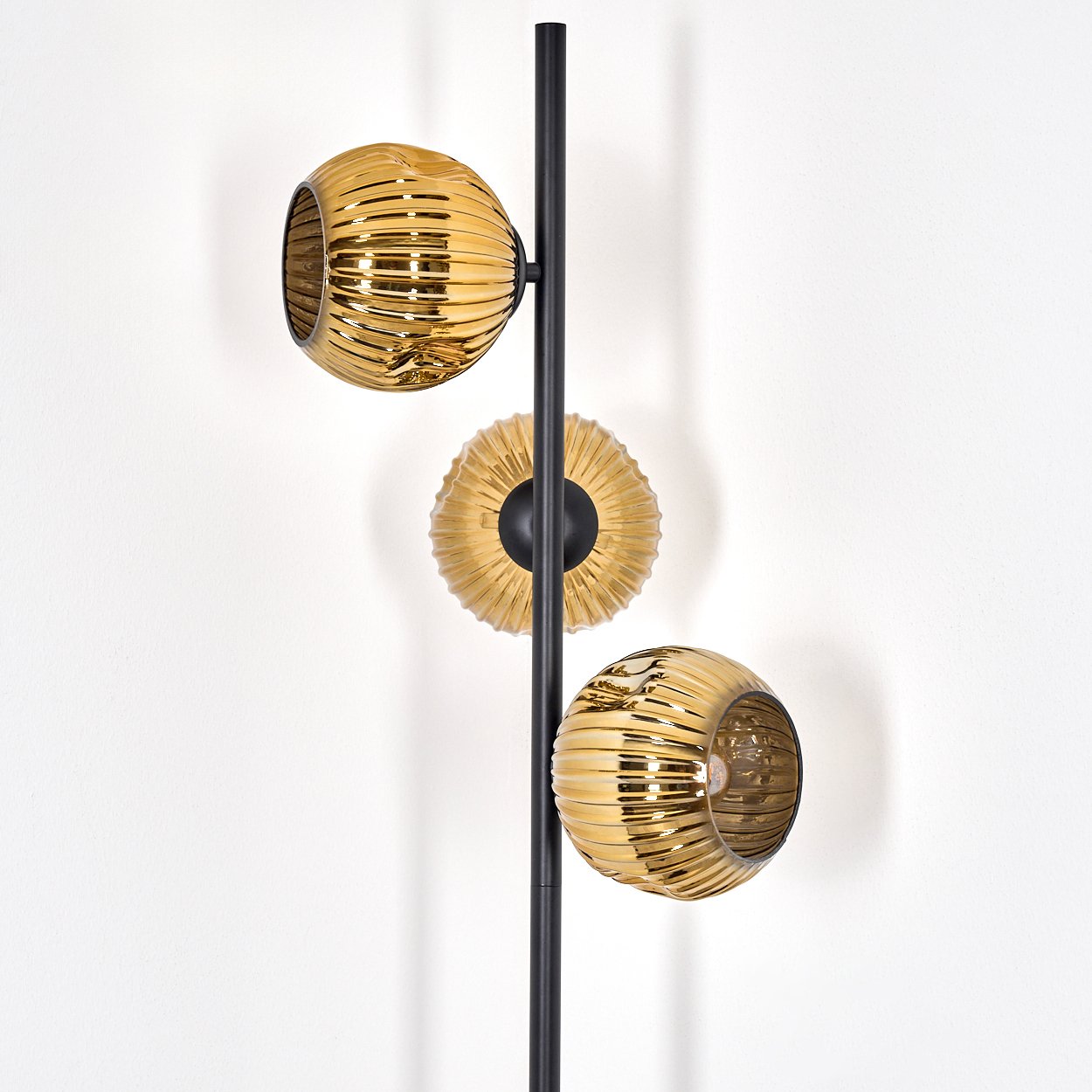 Eine moderne Lampe mit drei goldenen, geriffelten Kugeln, die an einer schwarzen Stange befestigt sind. Der Hintergrund ist eine weiße Wand.