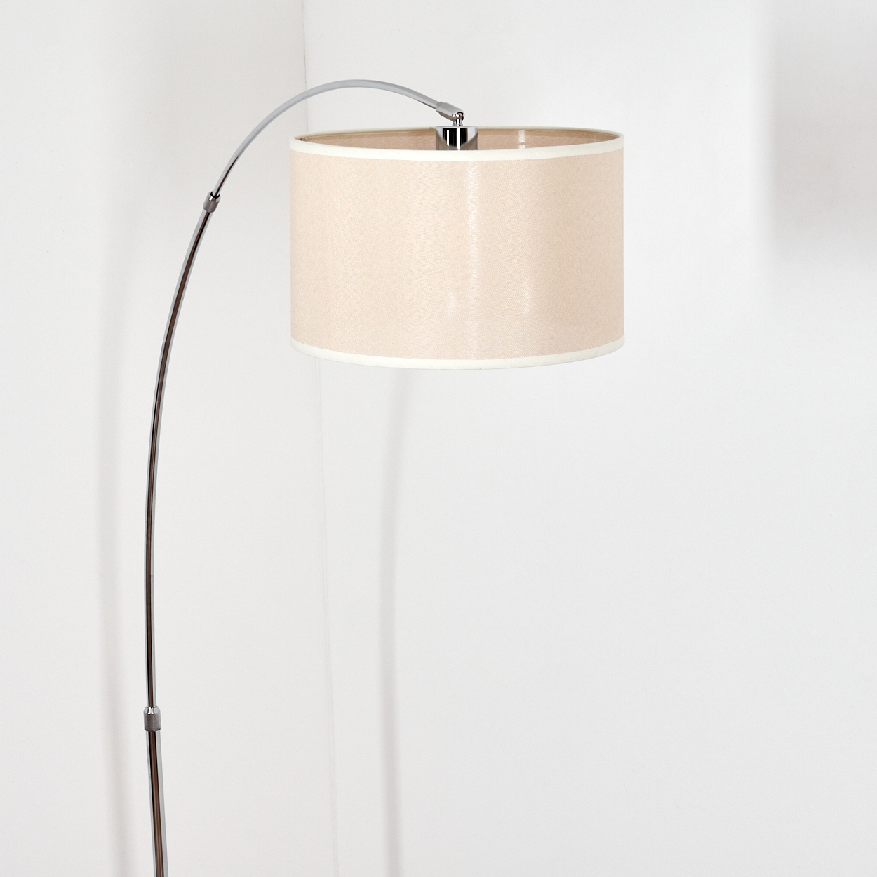 Eine moderne Bogen-Stehlampe mit beigem Lampenschirm. Die Lampe hat eine Chromoberfläche und steht vor einer weißen Wand.