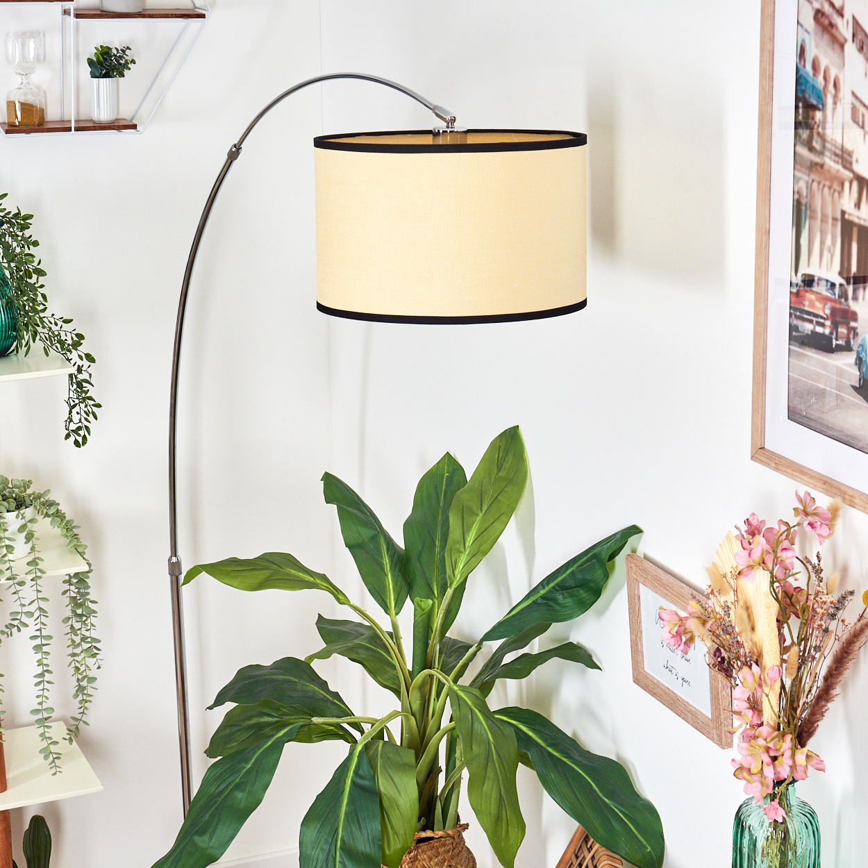 Eine Stehlampe mit beigem Schirm und schwarzem Rand, davor eine Pflanze in einem Korb. Regale und Bilder an der weißen Wand.