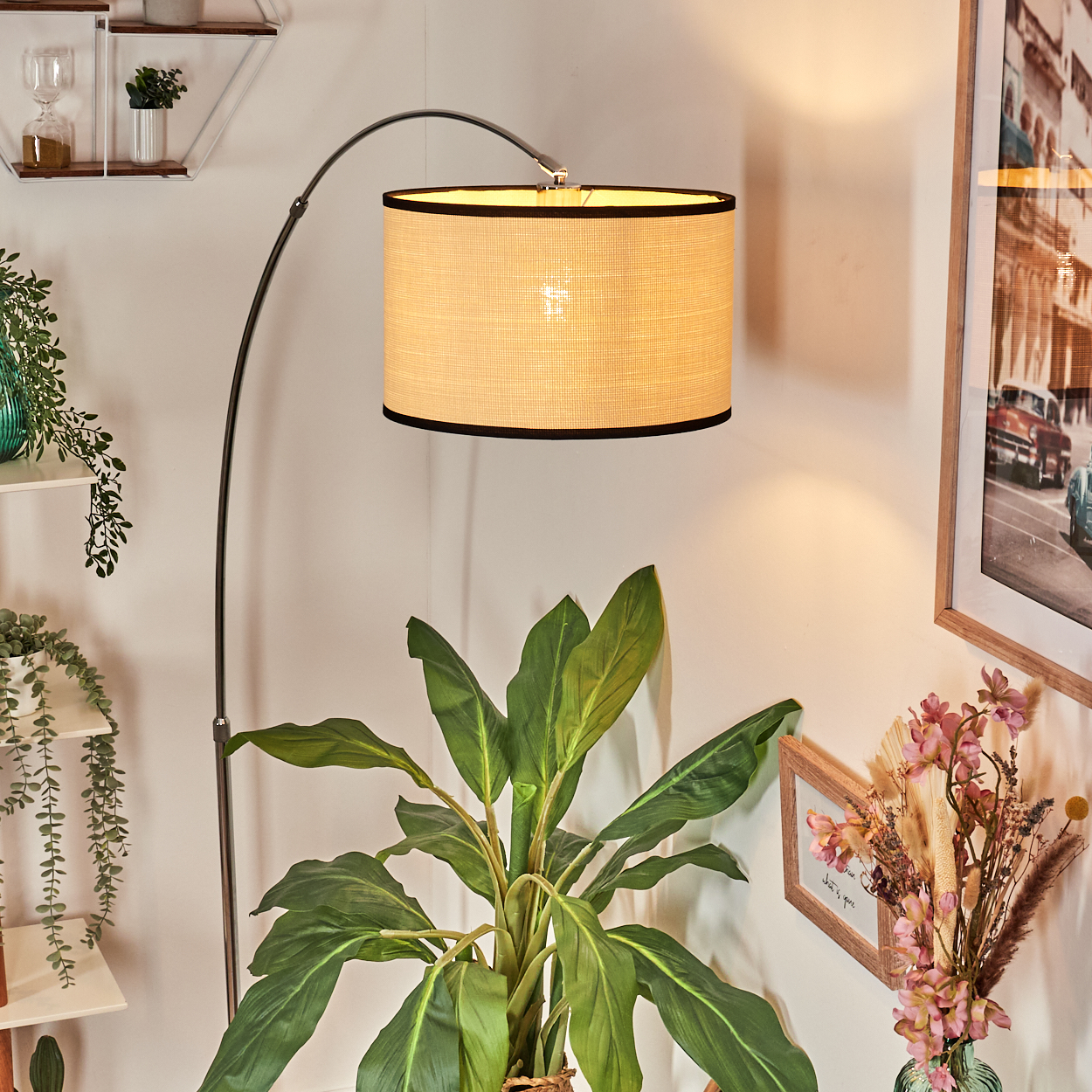 Eine Stehlampe mit beigem Lampenschirm. Davor eine große grüne Pflanze. Ein Bild und Blumen sind auf der rechten Seite.