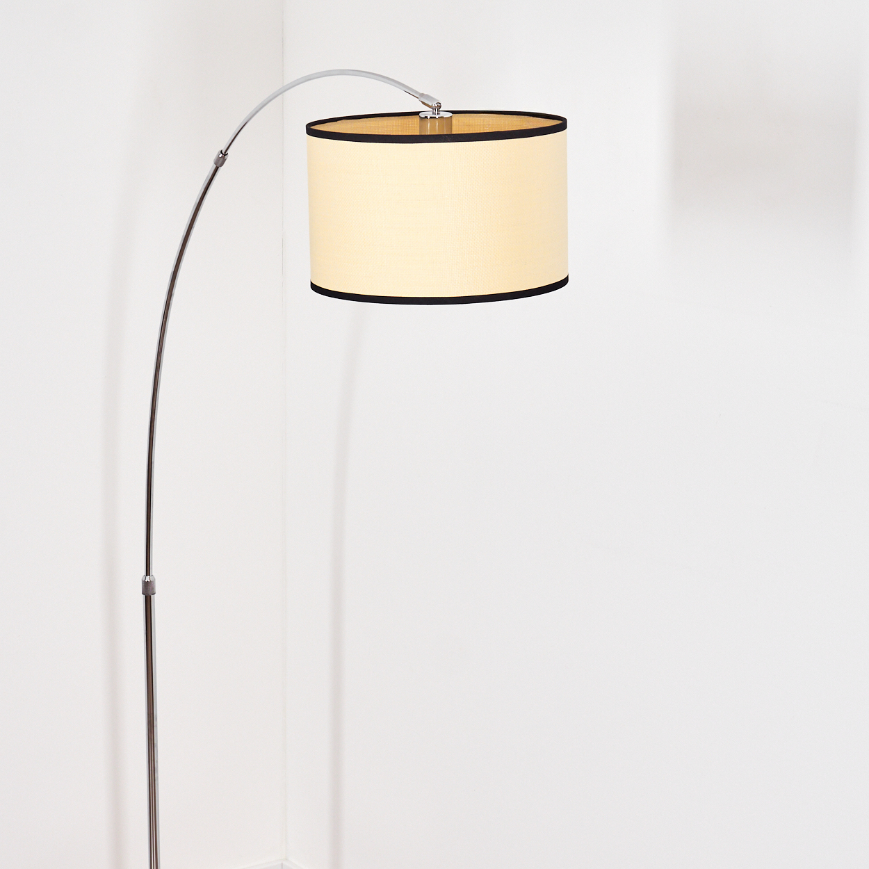 Eine moderne Bogenstehlampe mit beigefarbenem Lampenschirm und schwarzem Rand. Die Lampe steht vor einer weißen Wand in einer Ecke.
