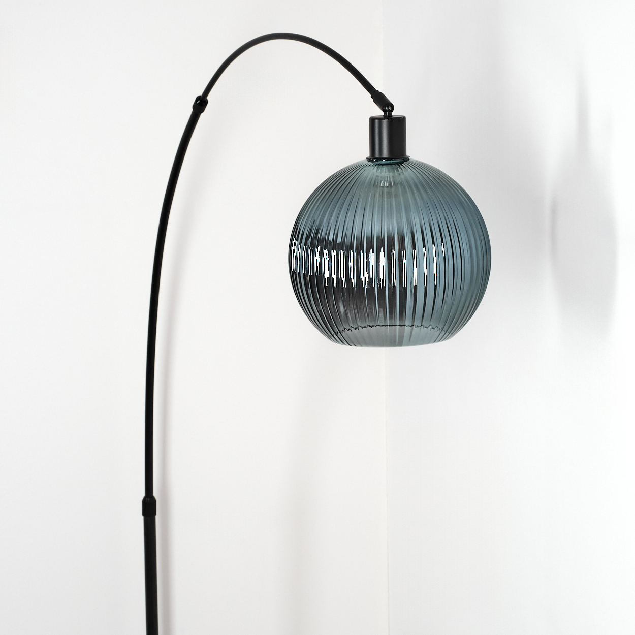 Eine moderne Lampe mit einem schwarzen, gebogenen Arm und einem runden, geriffelten, blau-grauen Glasschirm vor einer weißen Wand.