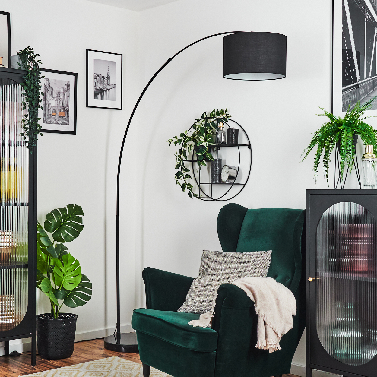 Ein grüner Sessel mit Decke und Kissen, eine schwarze Lampe, Topfpflanzen und gerahmte Bilder an einer weißen Wand.