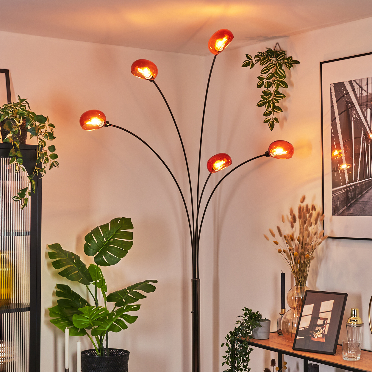 Eine Stehlampe mit roten Schirmen beleuchtet einen Raum mit Pflanzen, einem Bilderrahmen und einem Regal.