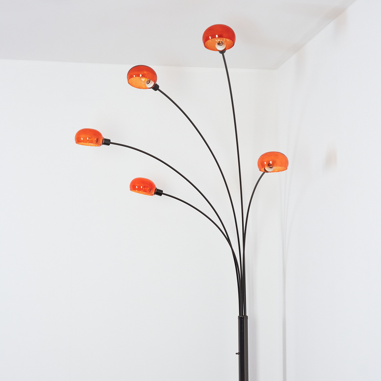 Eine moderne Stehlampe mit fünf orangefarbenen Glasschirmen an schwarzen Stielen. Die Lampe steht in einer weißen Ecke.