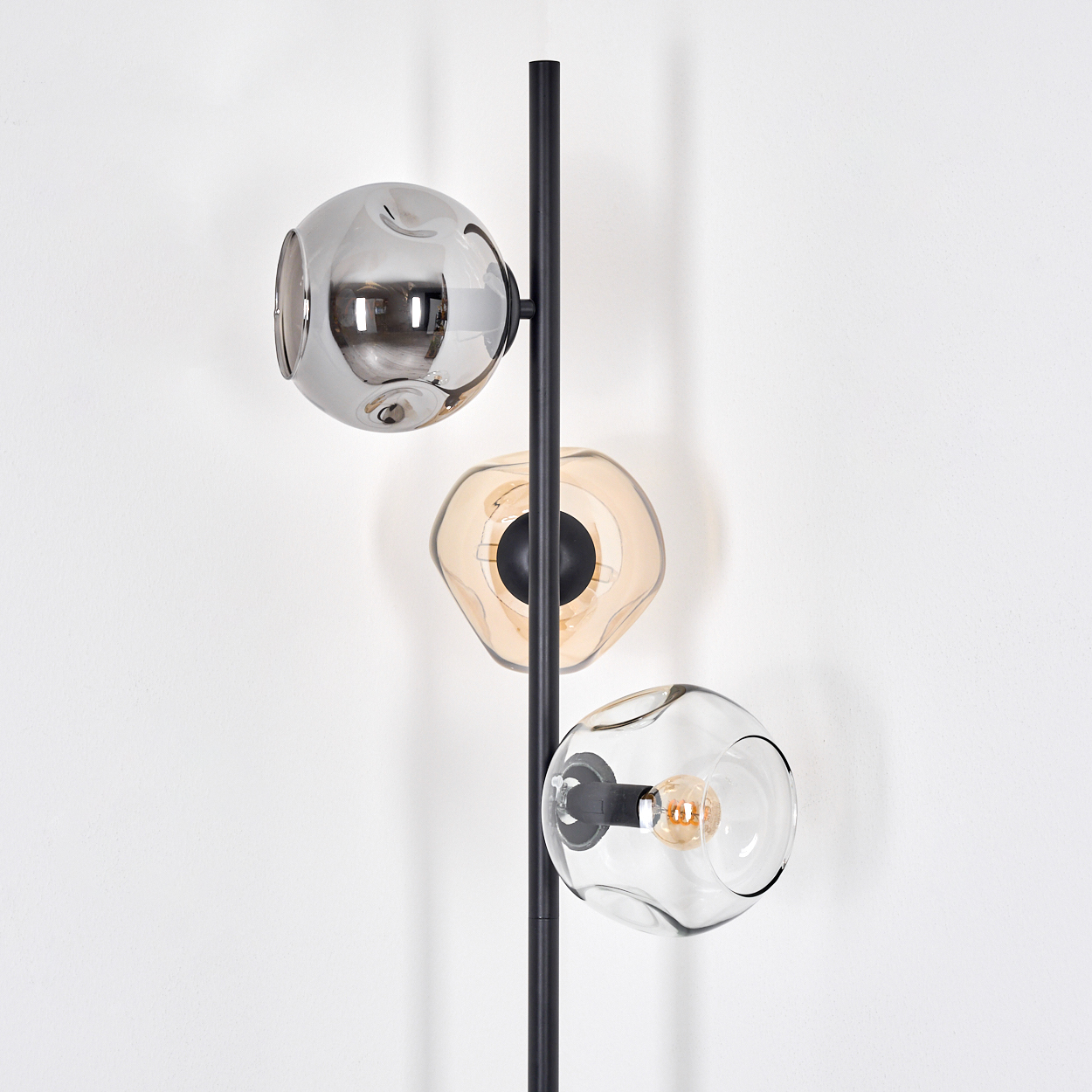 Eine moderne Stehlampe mit drei kugelförmigen Leuchten in verschiedenen Farben vor einer weißen Wand.