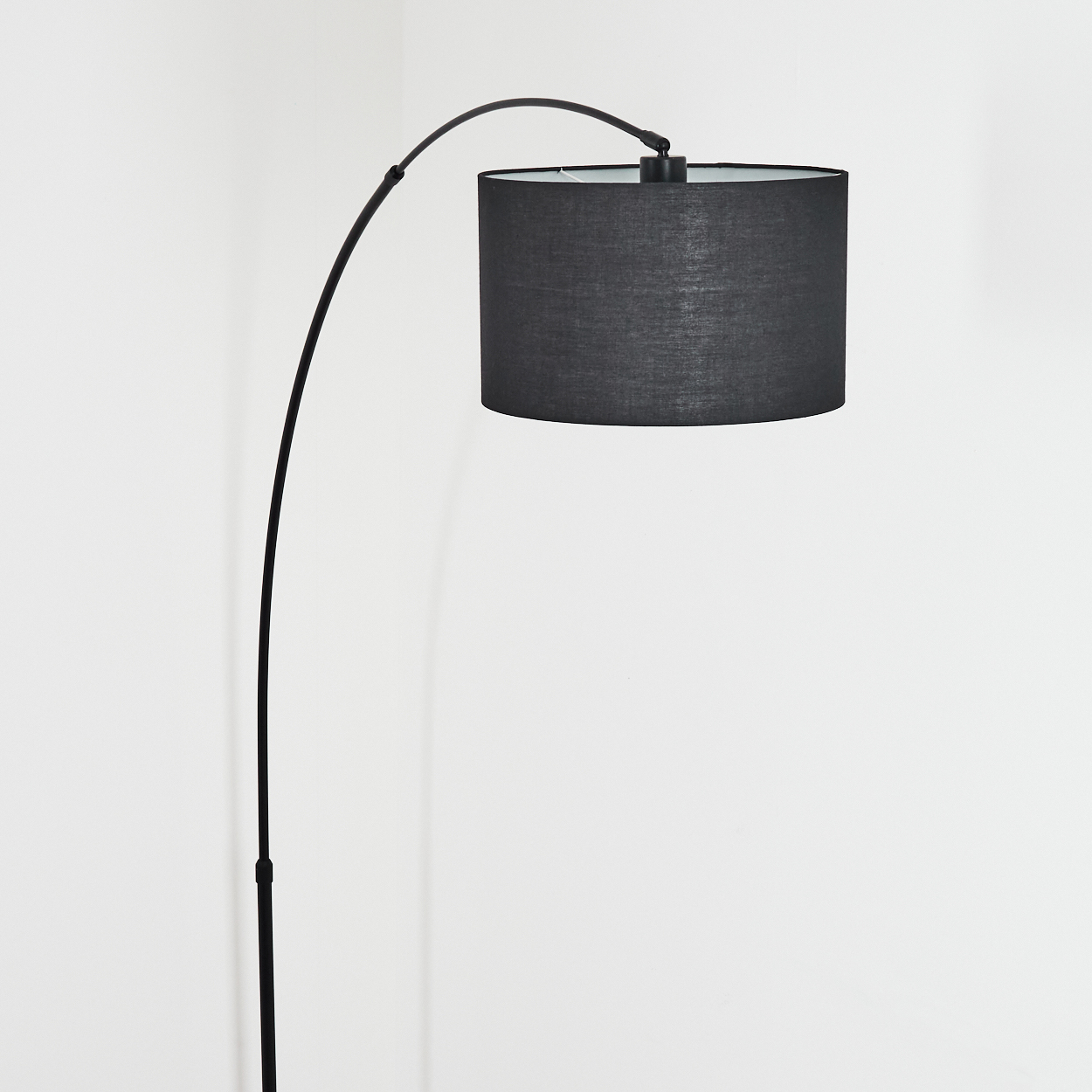 Eine schwarze Stehlampe mit gebogenem Arm und zylindrischem Lampenschirm vor weißer Wand.