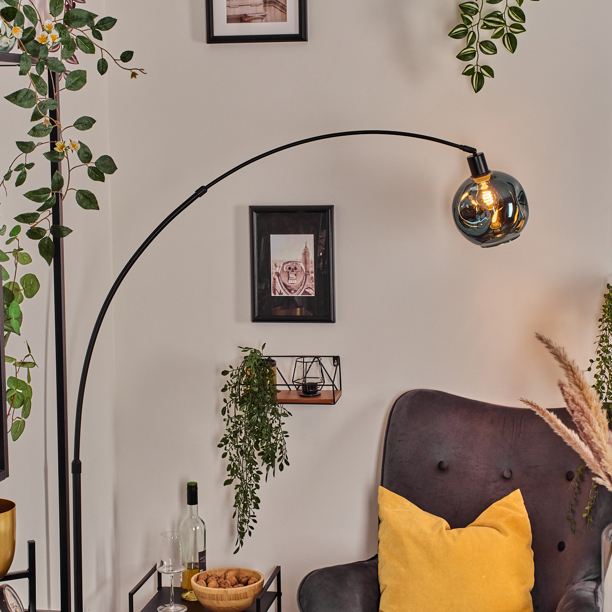 Eine Bogenlampe mit Glaskolben beleuchtet einen Raum mit einem grauen Sessel, einem gelben Kissen und Zierpflanzen.