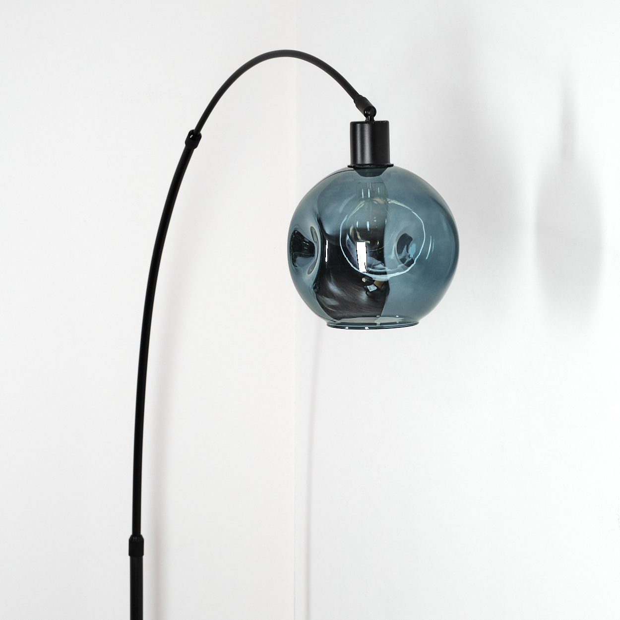 Eine moderne Lampe mit einem schwarzen, gebogenen Arm und einem blauen Glaskugel-Schirm steht in der Ecke. Im Hintergrund sind weiße Wände.