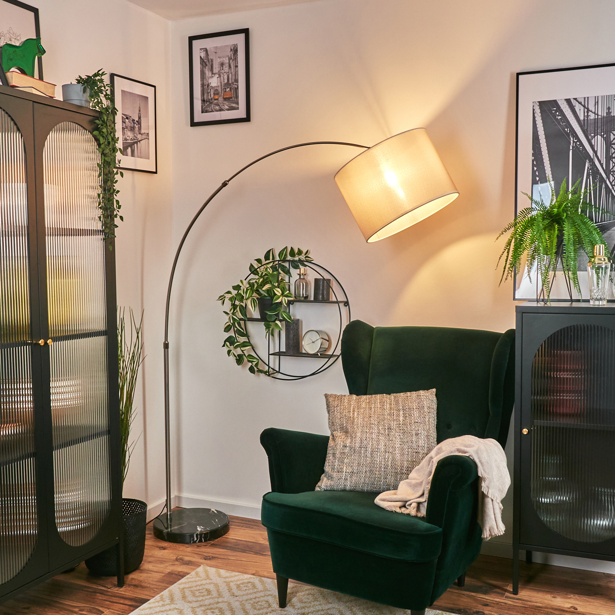 Ein grüner Sessel steht unter einer Lampe. Ein schwarzer Schrank und ein rundes Regal mit Pflanzen sind ebenfalls zu sehen. Bilder hängen an der Wand.
