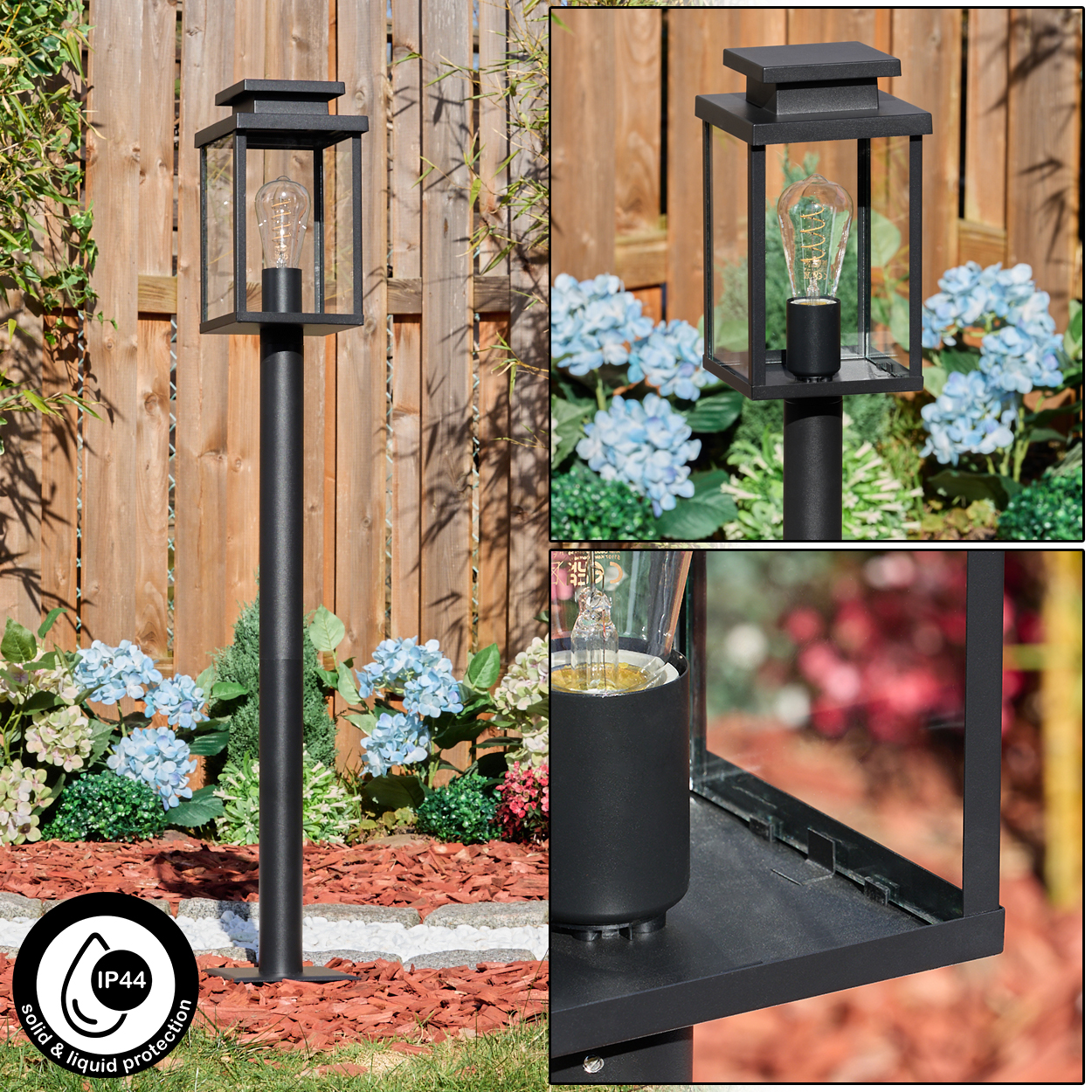 Eine schwarze Außenlampe mit Glasgehäuse. Der Hintergrund hat Blumen, Holzzaun und Mulch.