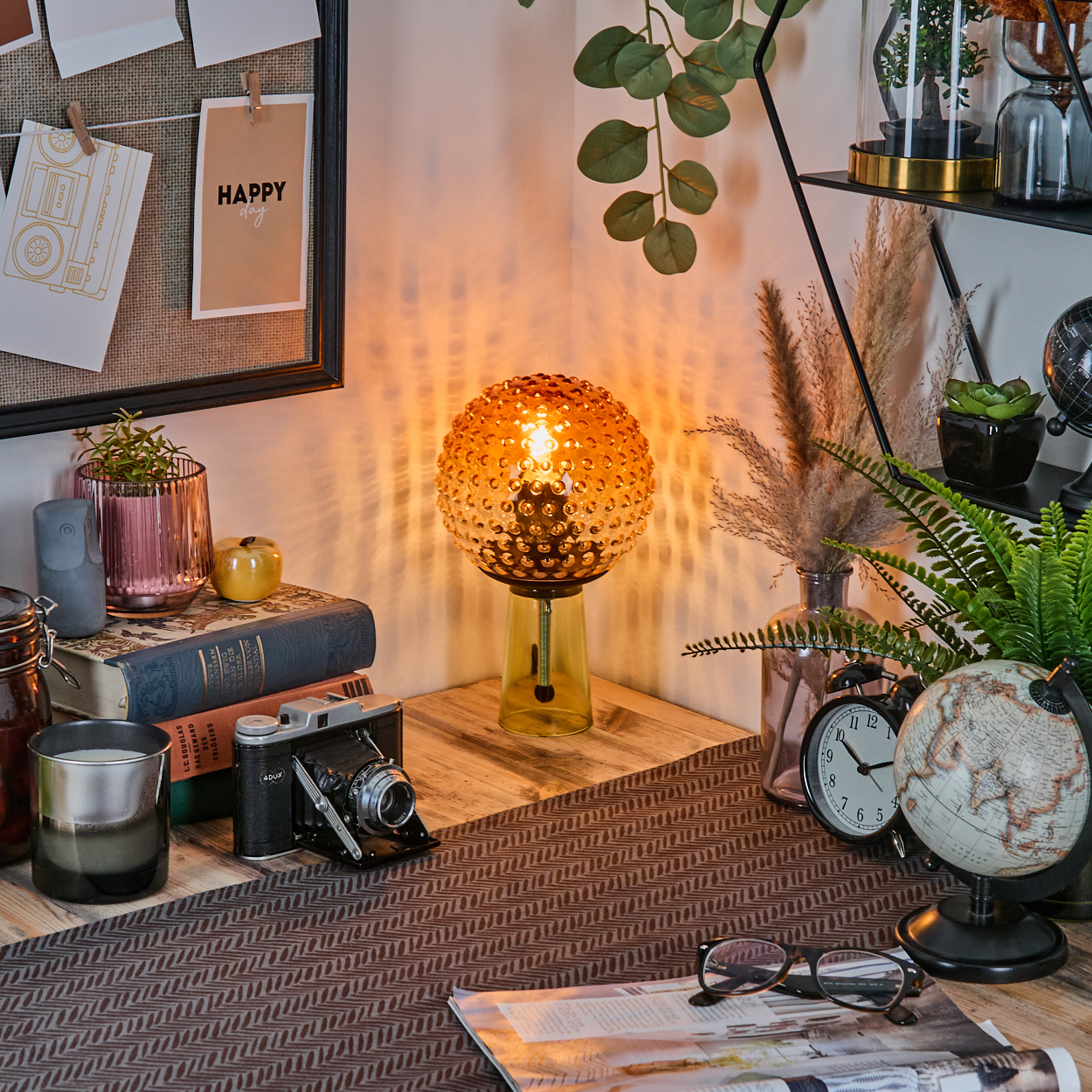 Ein Schreibtisch mit Lampe, Büchern, Kamera, Globus und anderen Dekorationsartikeln. Auf der Pinnwand steht eine Happy-Day-Nachricht.