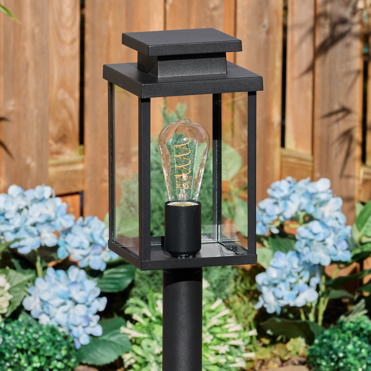 Eine schwarze Außenlampe mit Glasgehäuse und dekorativer Glühbirne steht im Fokus. Blaue Blumen und ein Holzzaun sind im Hintergrund.