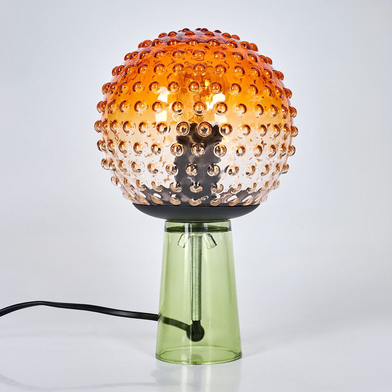 Eine dekorative Lampe mit einem orange-braunen, strukturierten Globus-Schirm und einem grünen Glassockel. Ein schwarzes Kabel ist angeschlossen.