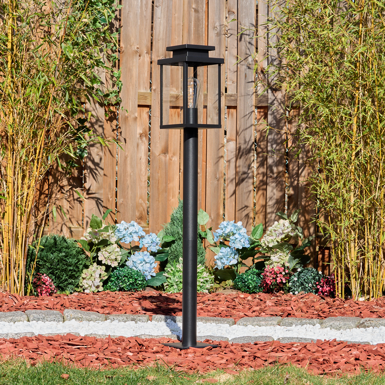 Schwarze Außenlampe in einem Garten mit Bambus, blauen Hortensien und Holzhackschnitzelmulch. Ein Holzzaun ist im Hintergrund.