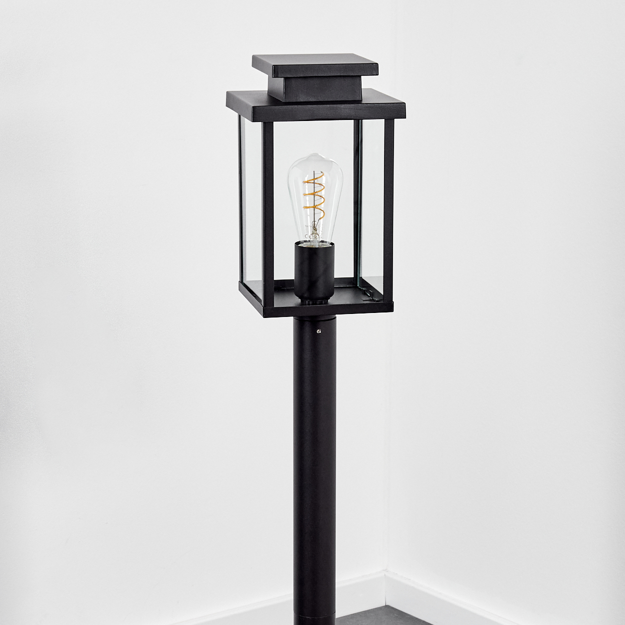 Eine schwarze Außenlampe mit Glasgehäuse und sichtbarer Glühbirne steht vor einer weißen Wand. Die Lampe steht auf einem schwarzen Pfosten.