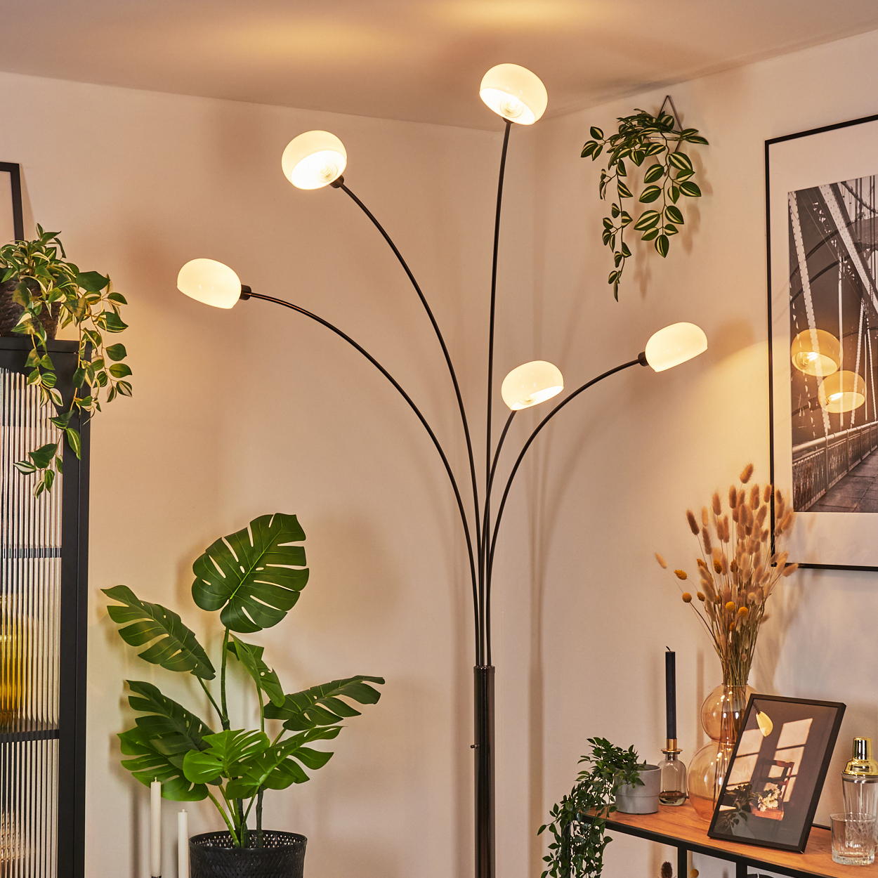 Eine moderne Stehlampe mit fünf Lichtern beleuchtet einen Raum. Pflanzen, ein Bild und ein Holzregal sind im Bild.