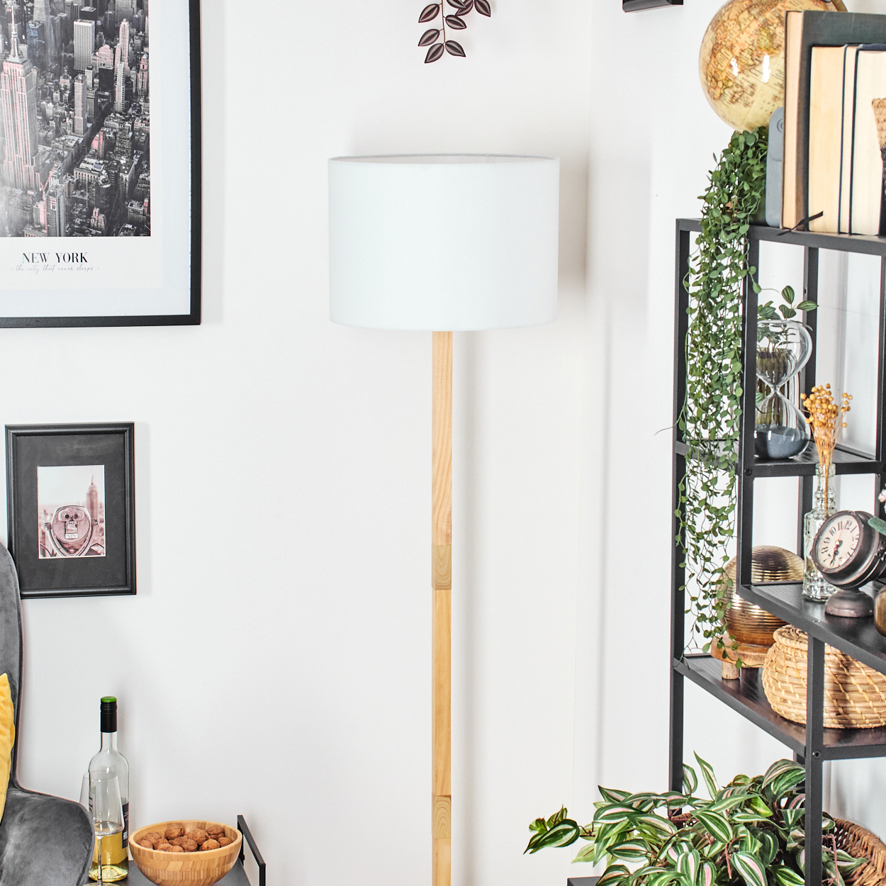 Ein Raum mit einer Stehlampe. Es gibt ein Regal mit Dekoration, ein New York-Poster, ein gerahmtes Bild, einen Tisch mit Nüssen und Wein.