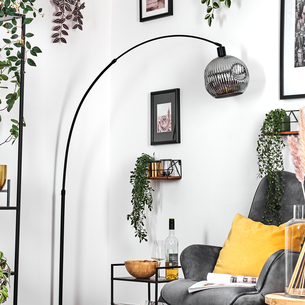 Ein modernes Wohnzimmer mit schwarzer Bogenlampe, grauem Sessel, gelbem Kissen und Zierpflanzen. Eine Weinflasche und ein Glas stehen auf dem Beistelltisch.