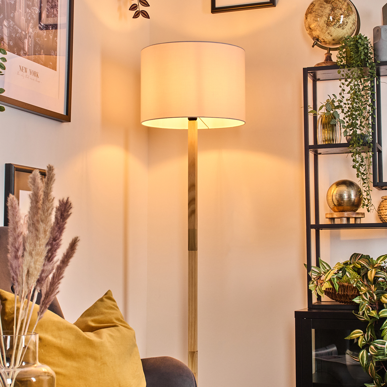 Eine Stehlampe steht neben einer Wand. Rechts ein Regal mit Pflanzen und Globus. Links eine Vase mit Pampasgras.