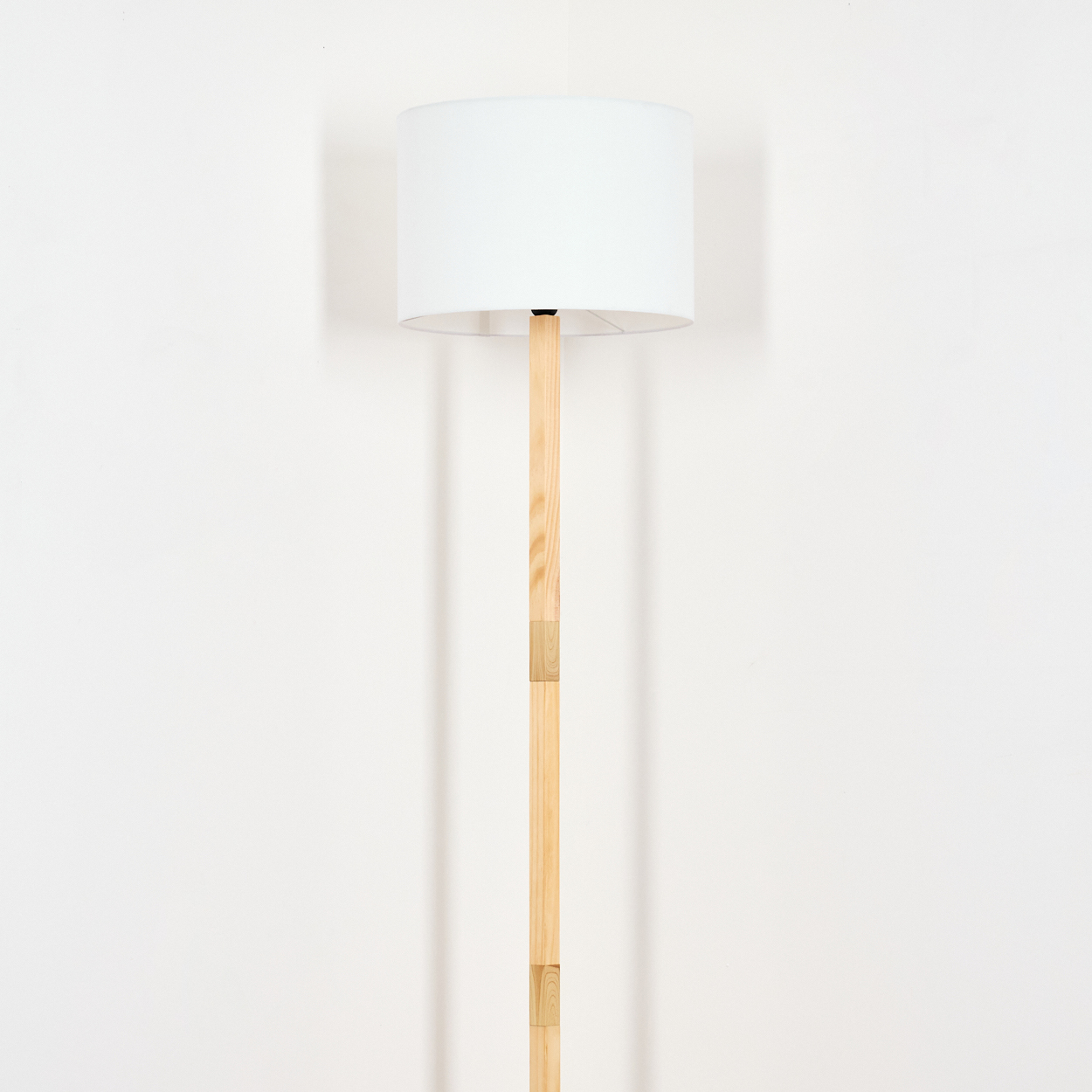 Eine Stehlampe mit weißem Lampenschirm und Holzstange steht vor einer weißen Wand. Schatten der Lampe sind an der Wand.