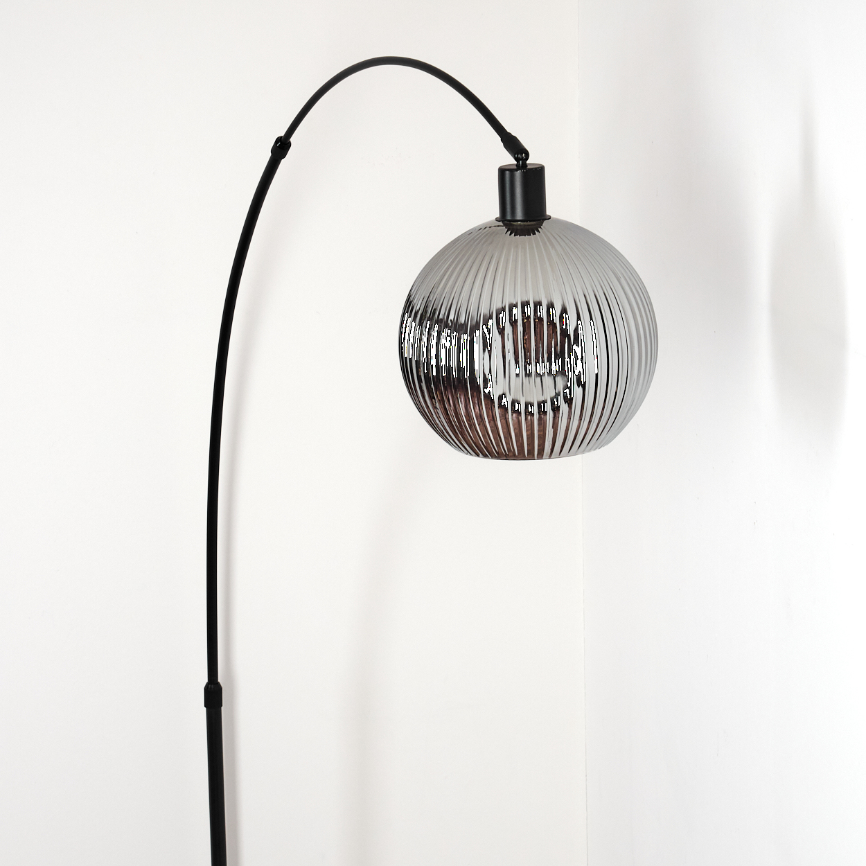 Eine moderne Stehlampe mit gebogenem schwarzem Stiel und einem runden, geriffelten, silbernen Lampenschirm vor weißer Wand.