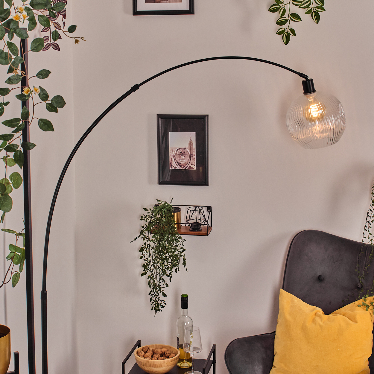 Ein Raum mit einer schwarzen Bogenlampe, einem grauen Sessel mit gelbem Kissen und einem Tisch mit Snacks und Wein. Grüne Pflanzen sind zu sehen.