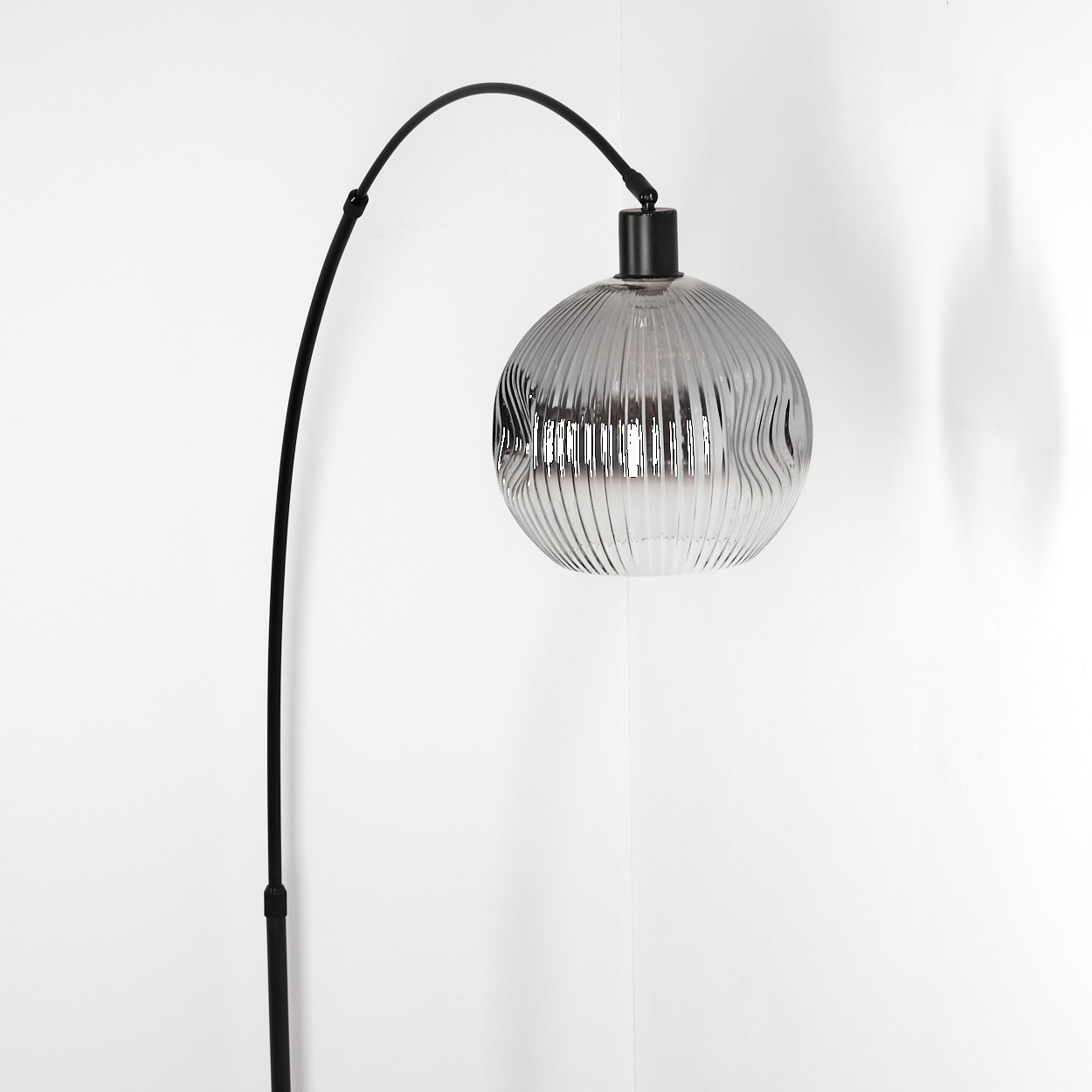 Eine moderne schwarze Stehlampe mit gebogenem Arm und geripptem, transparentem Glasschirm vor weißer Wand.
