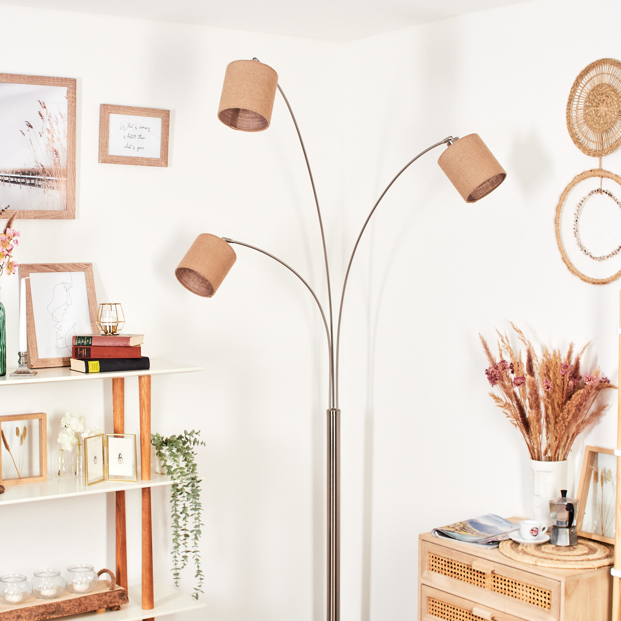 Eine Stehlampe mit drei Lampenschirmen steht in einem Raum. Es gibt ein Regal mit Büchern und eine Vase mit Blumen. Wanddekoration ist sichtbar.