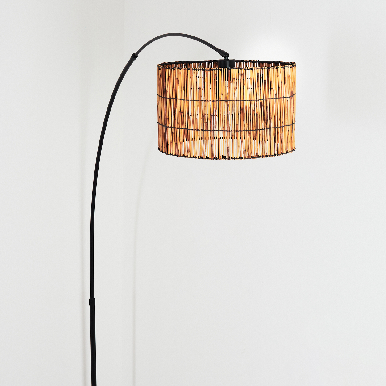 Eine Stehlampe mit schwarzem, gebogenem Arm und einem zylindrischen Lampenschirm aus Bambusstäben. Der Hintergrund ist eine weiße Wand.