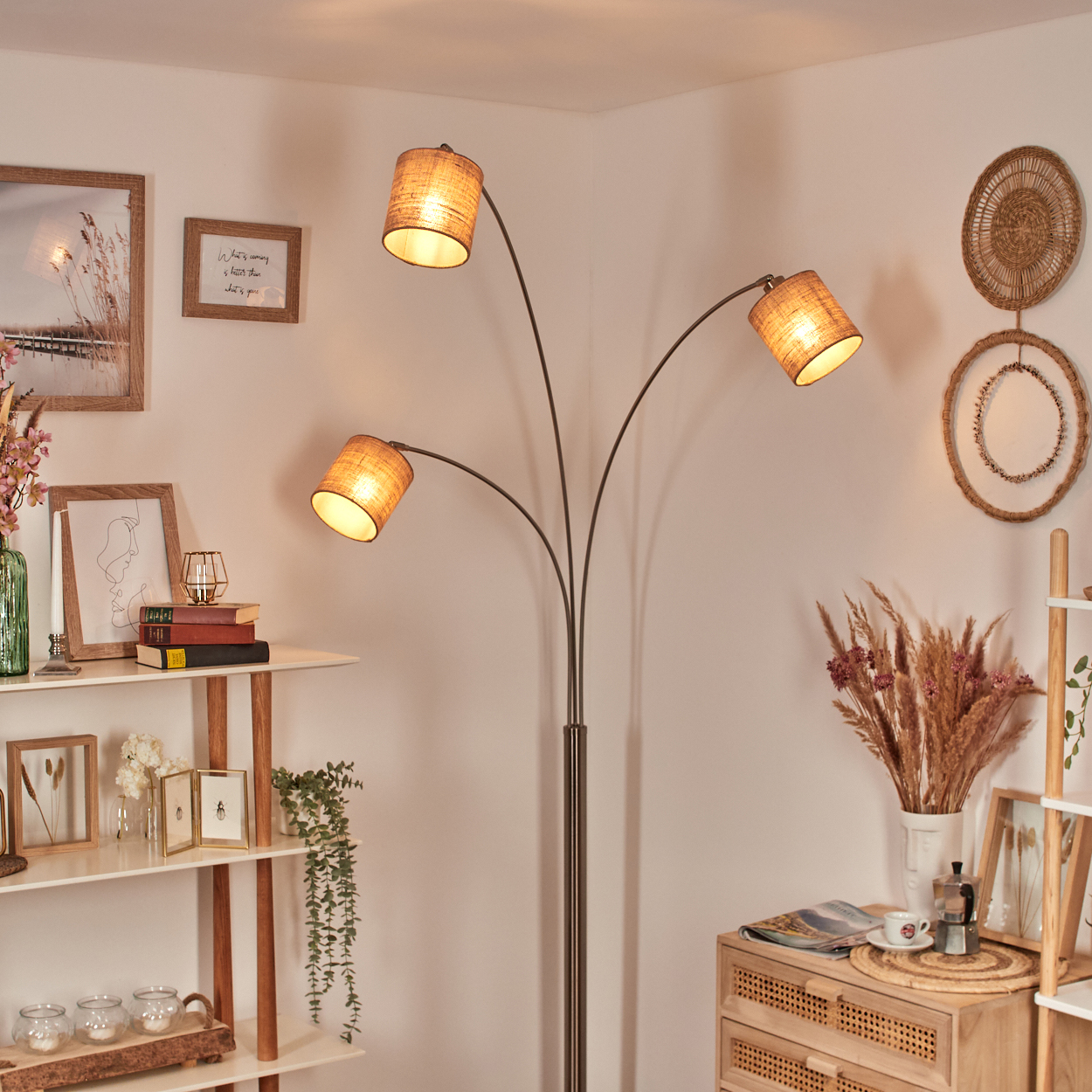 Eine Stehlampe mit drei Lampenschirmen beleuchtet einen gemütlichen Raum. Regale zeigen Dekorationen, darunter Bilder, Bücher und Kerzen. Wanddekoration.
