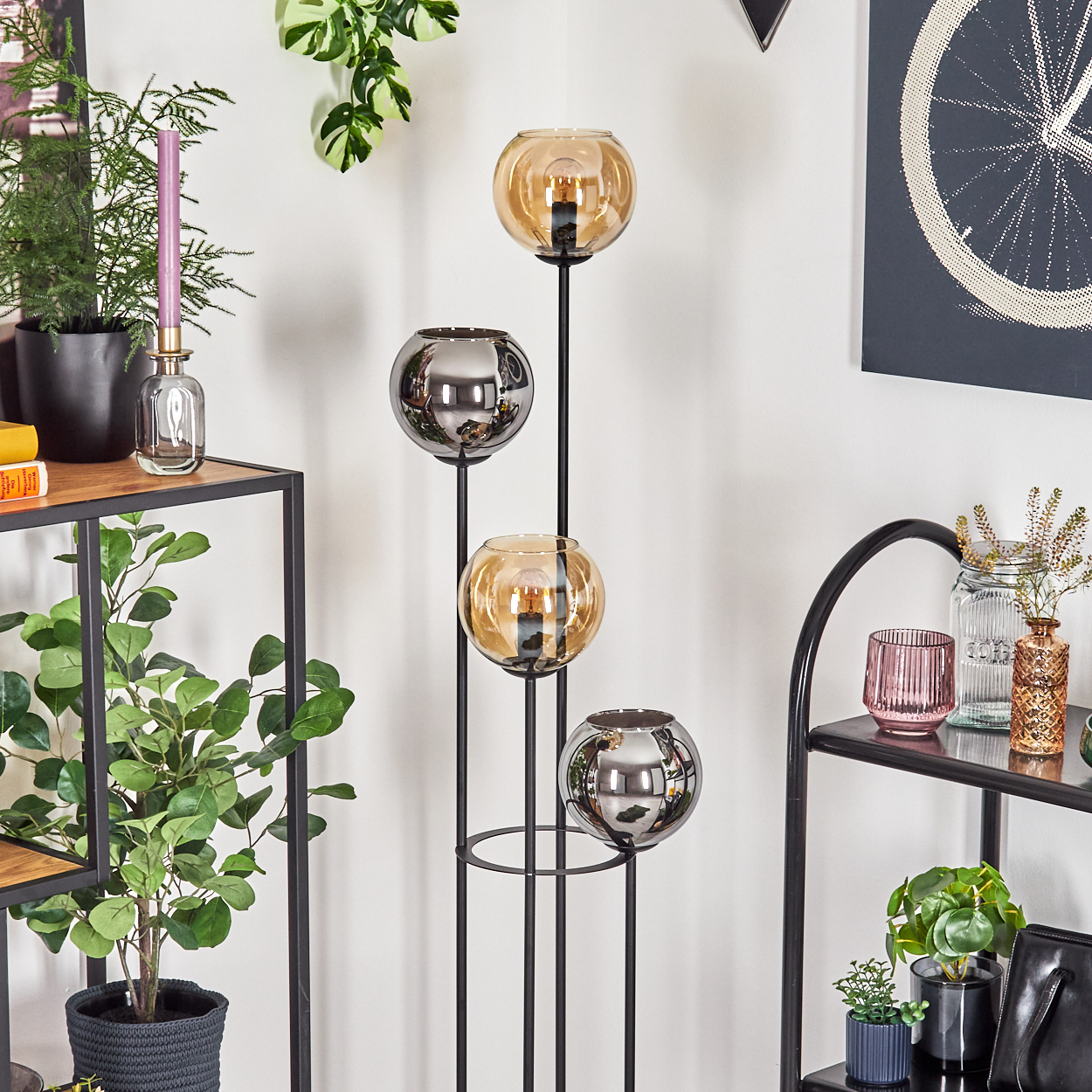 Eine dekorative Lampe mit vier Glaskugeln, Pflanzen und einem Fahrradrad-Kunstwerk werden vor einer weißen Wand gezeigt.