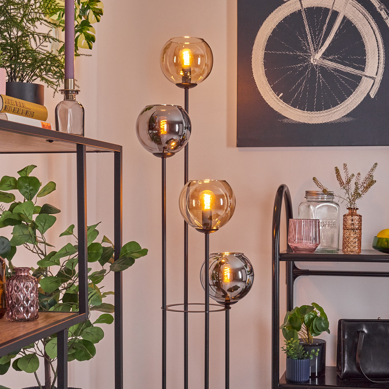 Ein moderner Wohnraum mit einer hohen Lampe mit vier Kugellampen. Regale halten Pflanzen, Bücher und Dekorationsartikel. Fahrradrad-Kunst hängt.