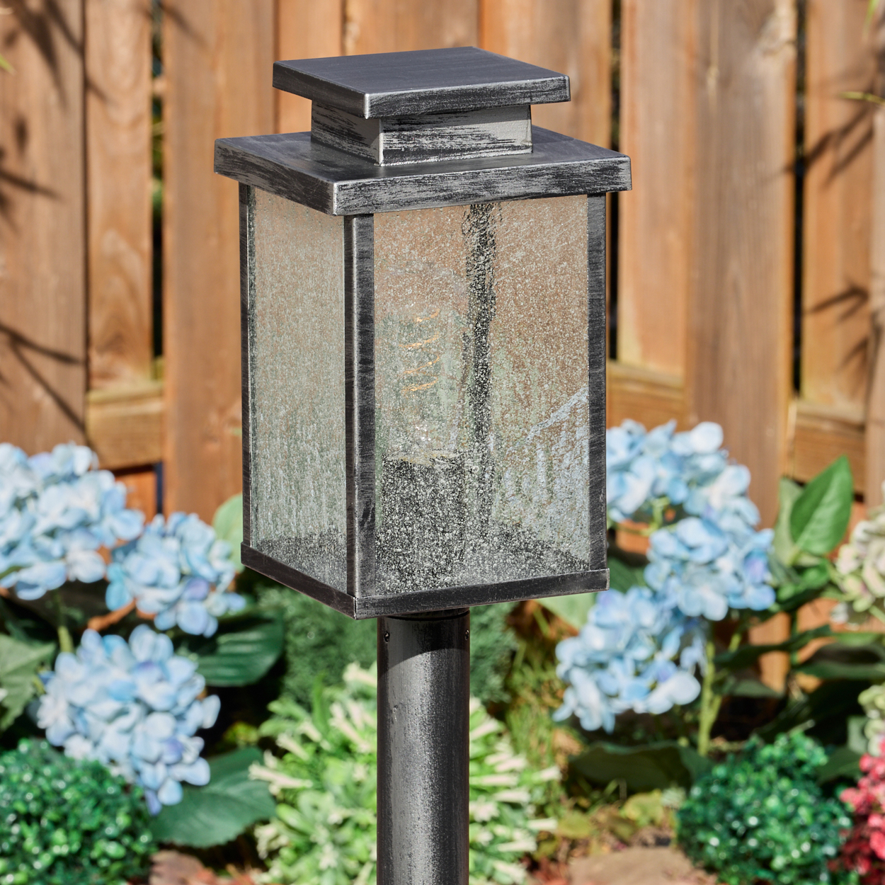 Eine dekorative Außenlampe mit quadratischem Design. Sie hat einen schwarzen Metallrahmen und Milchglasscheiben. Blaue Blumen sind im Hintergrund.